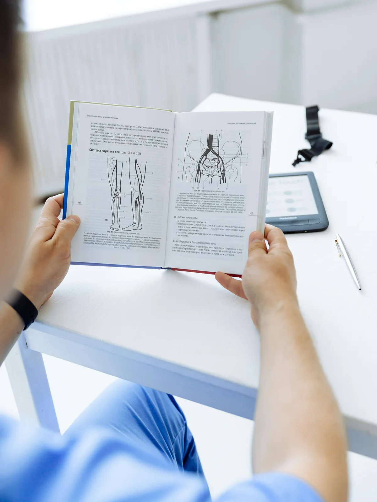 Healthcare professional in blue scrubs reading a medical anatomy textbook at a desk with a digital reader nearby