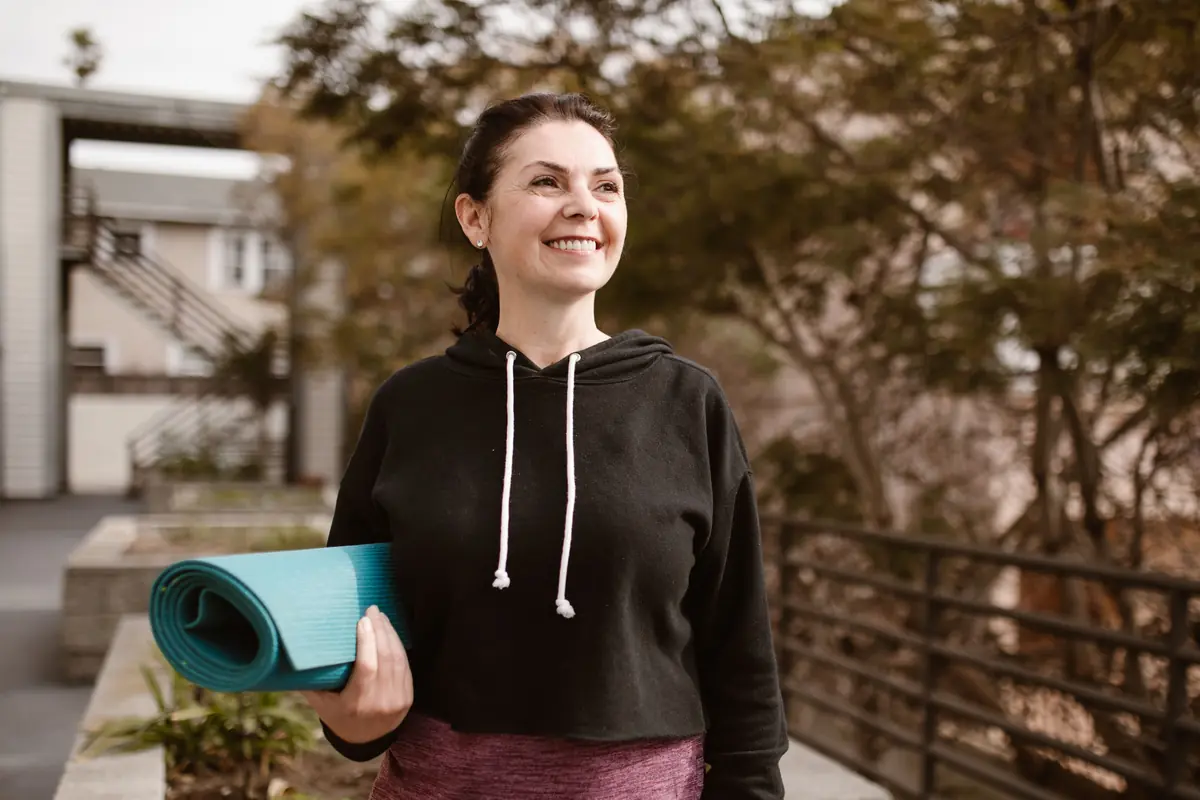 Woman smiling and holding a yoga mat outdoors after exercise, illustrating the mood and wellbeing benefits of regular physical activity
