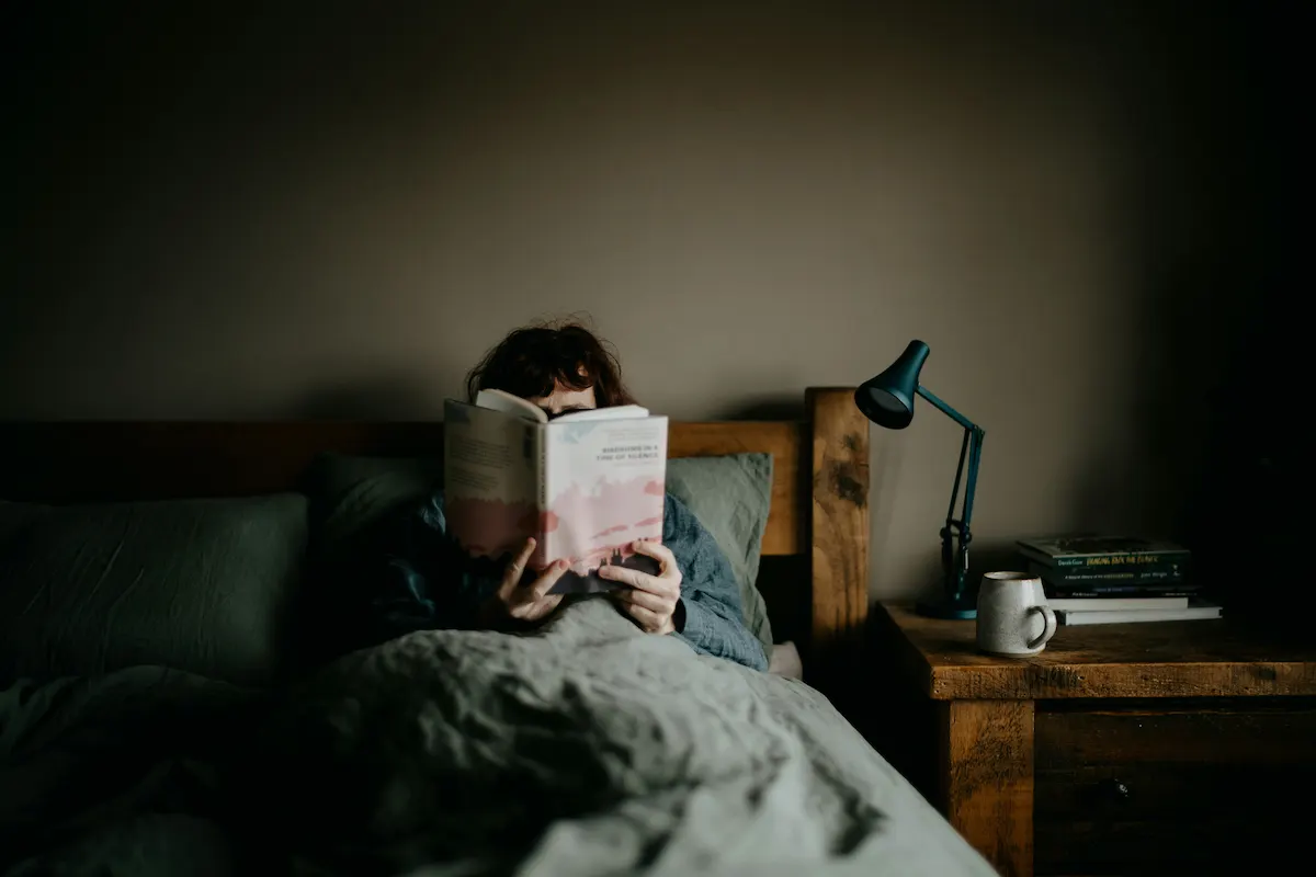 Person reading a physical book in bed with warm lamp light, a healthy wind-down routine before sleep