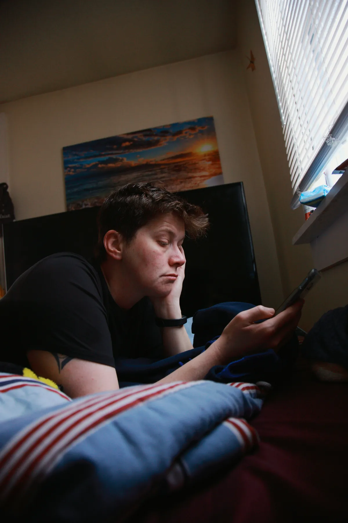 Teenager lying alone in a dim bedroom looking at a smartphone, looking troubled