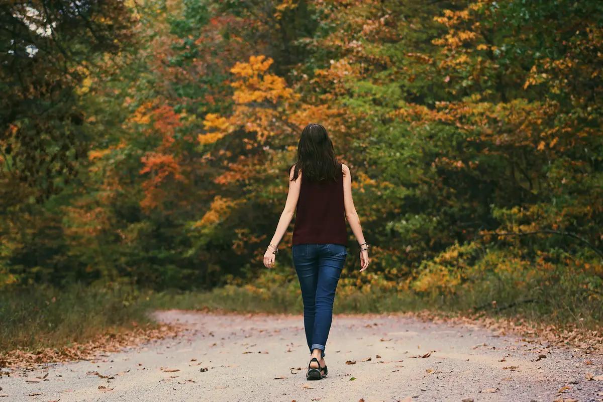 Woman walking alone on a forest trail surrounded by autumn foliage, representing the stress-reducing effects of nature exposure