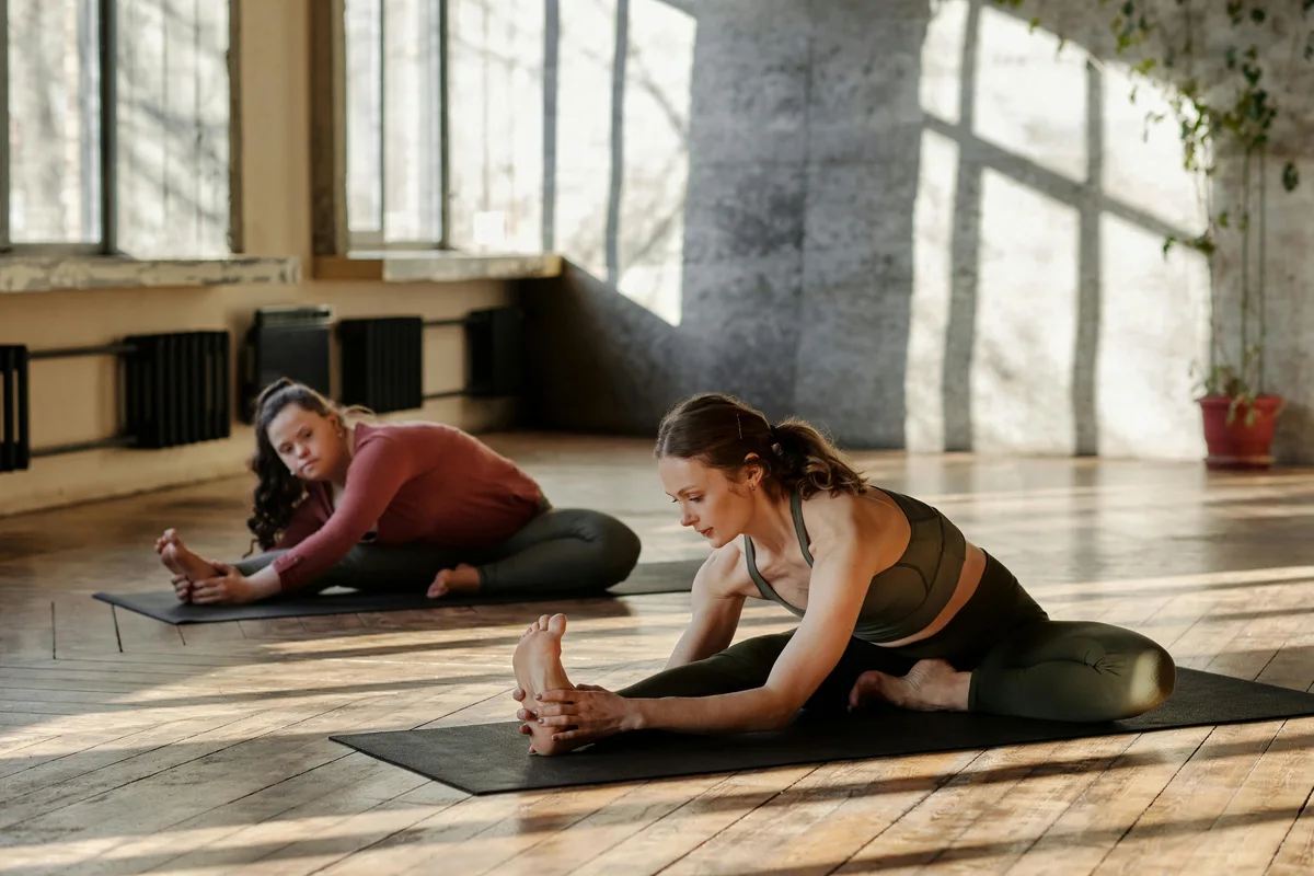Two women performing static forward stretches on mats in a studio with natural light